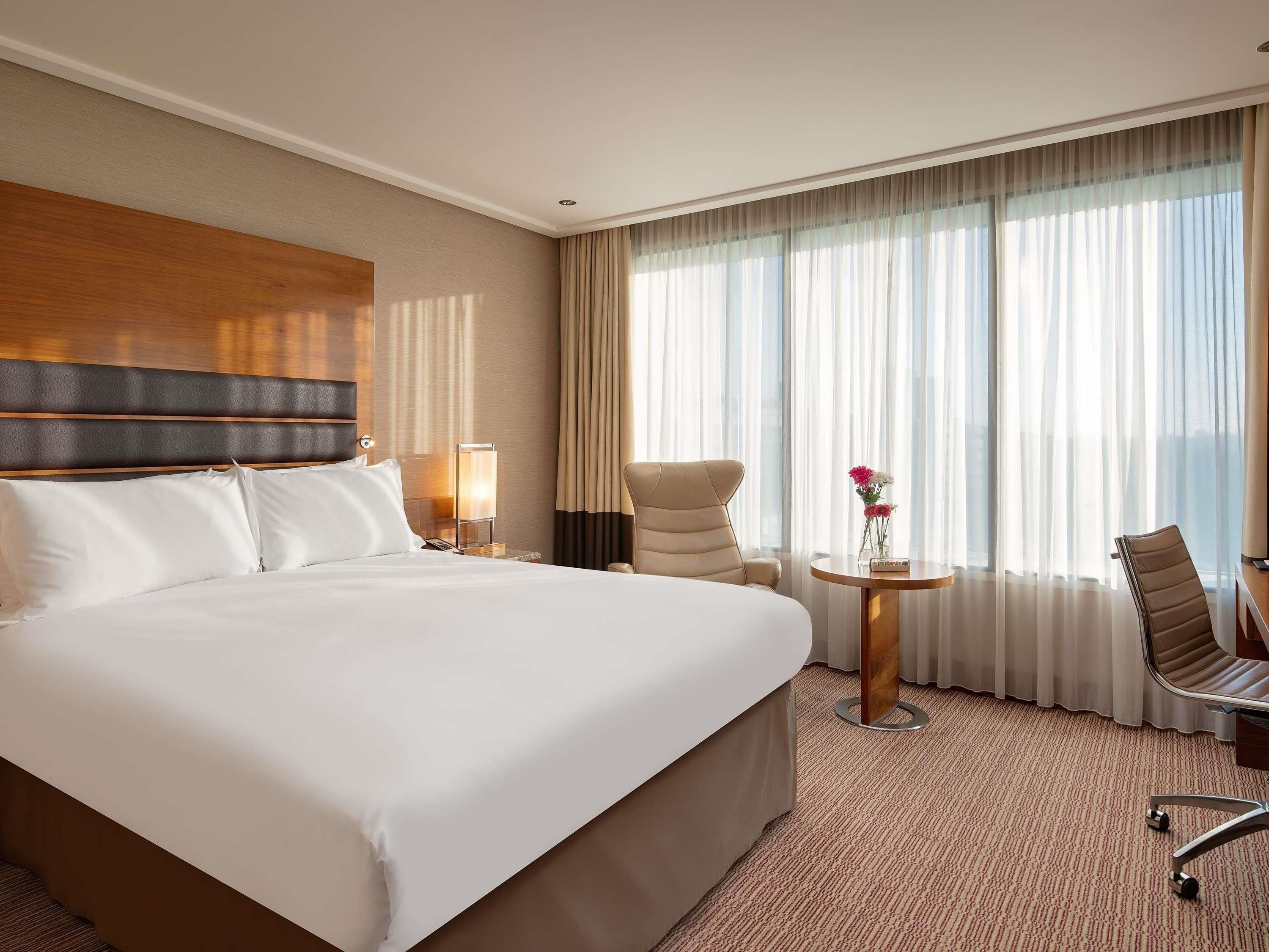 A hotel room with a large bed, a desk with a chair, a small table with flowers, and a view of the city through sheer curtains.
