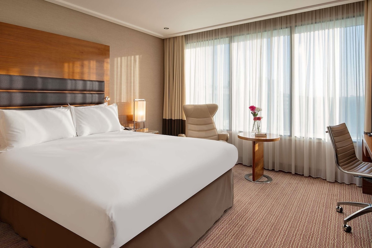 A hotel room with a large bed, a desk with a chair, a small table with flowers, and a view of the city through sheer curtains.