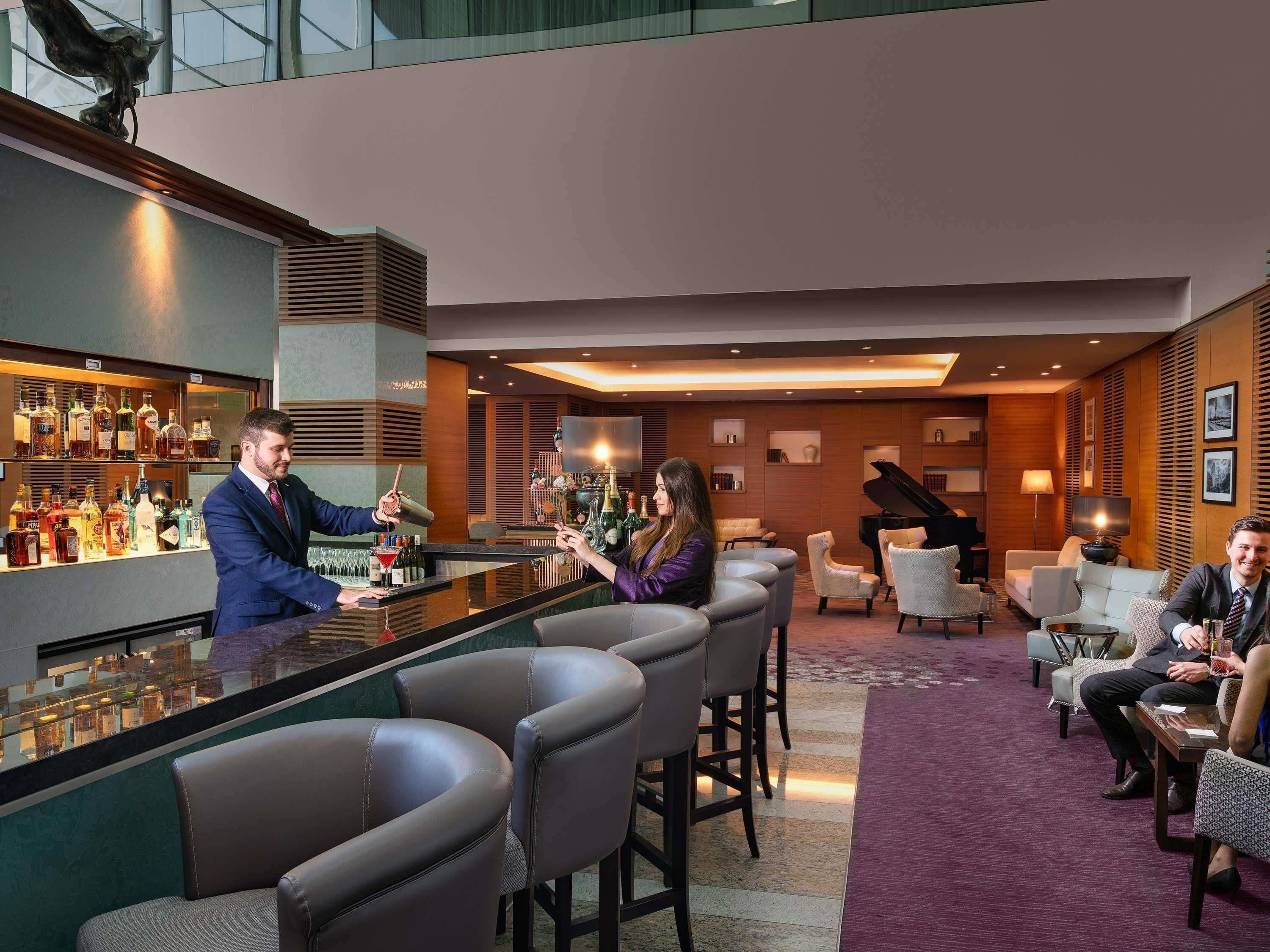 A bar area with a bartender, a seated woman, and a seated man. There are multiple chairs and a piano in the background.