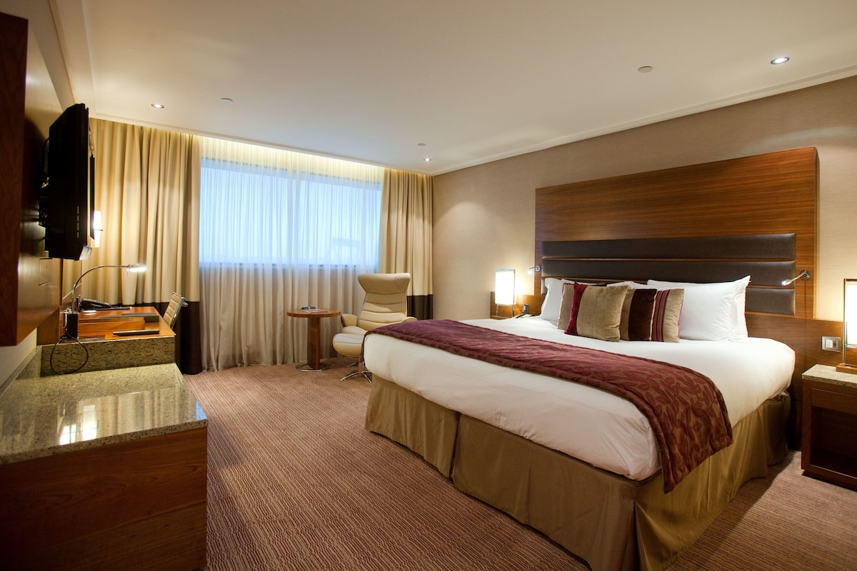 A hotel room with a large bed, a desk with a lamp, a chair, a TV, and a window with curtains.