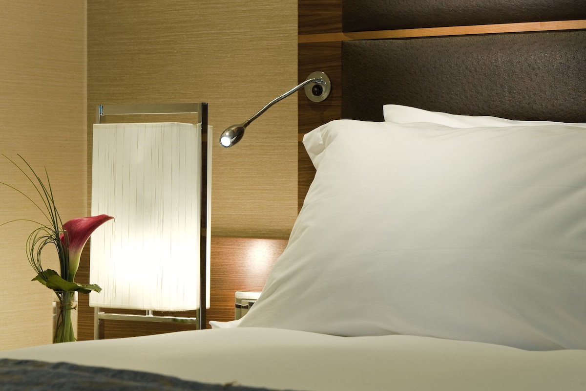 A hotel room with a bed, a bedside lamp, and a vase with a flower.