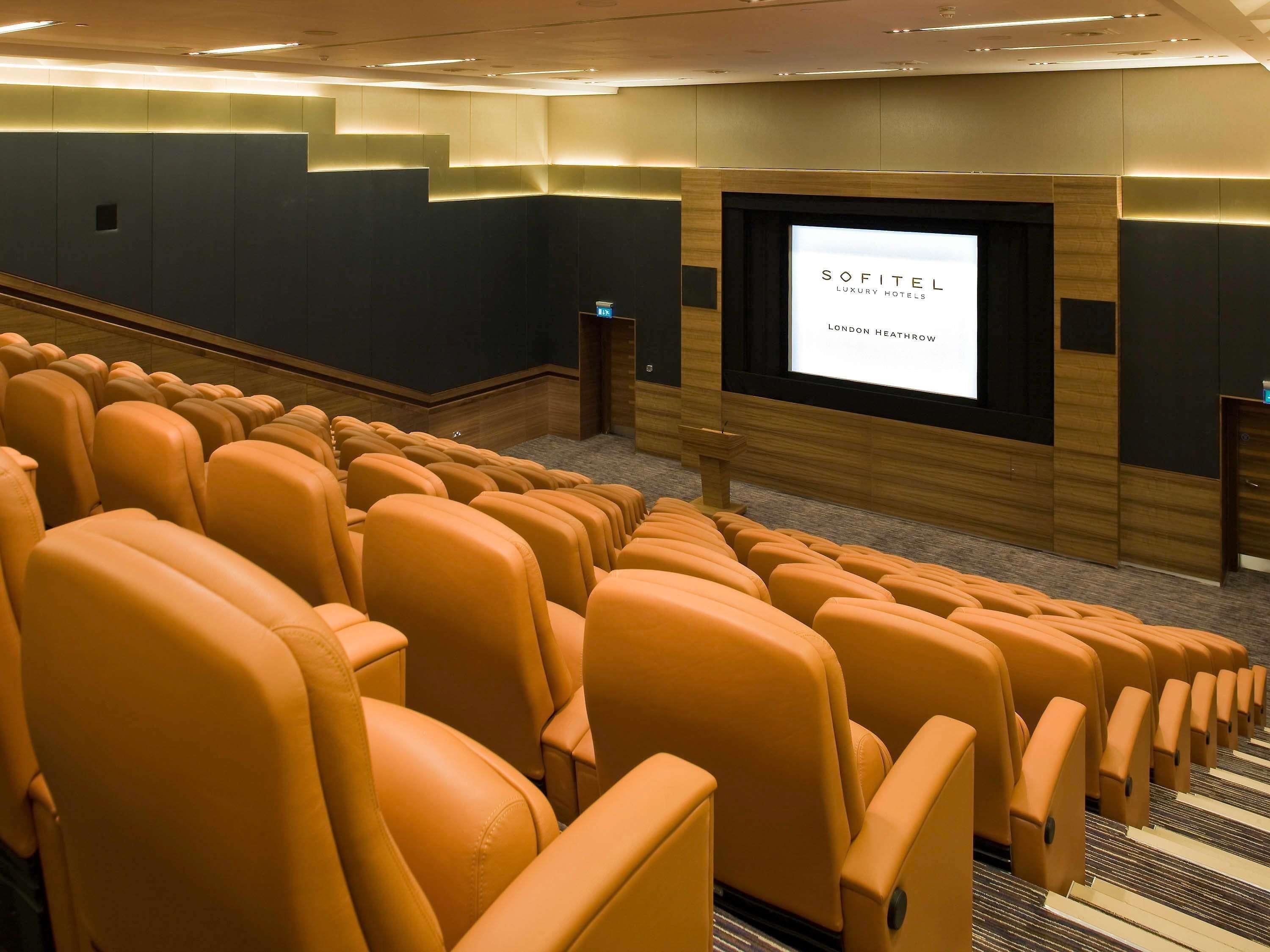 Un cine con filas de asientos naranja, una pantalla mostrando 'SOFITEL LUXURY HOTELS LONDON HEATHROW' y una pared de paneles de madera.