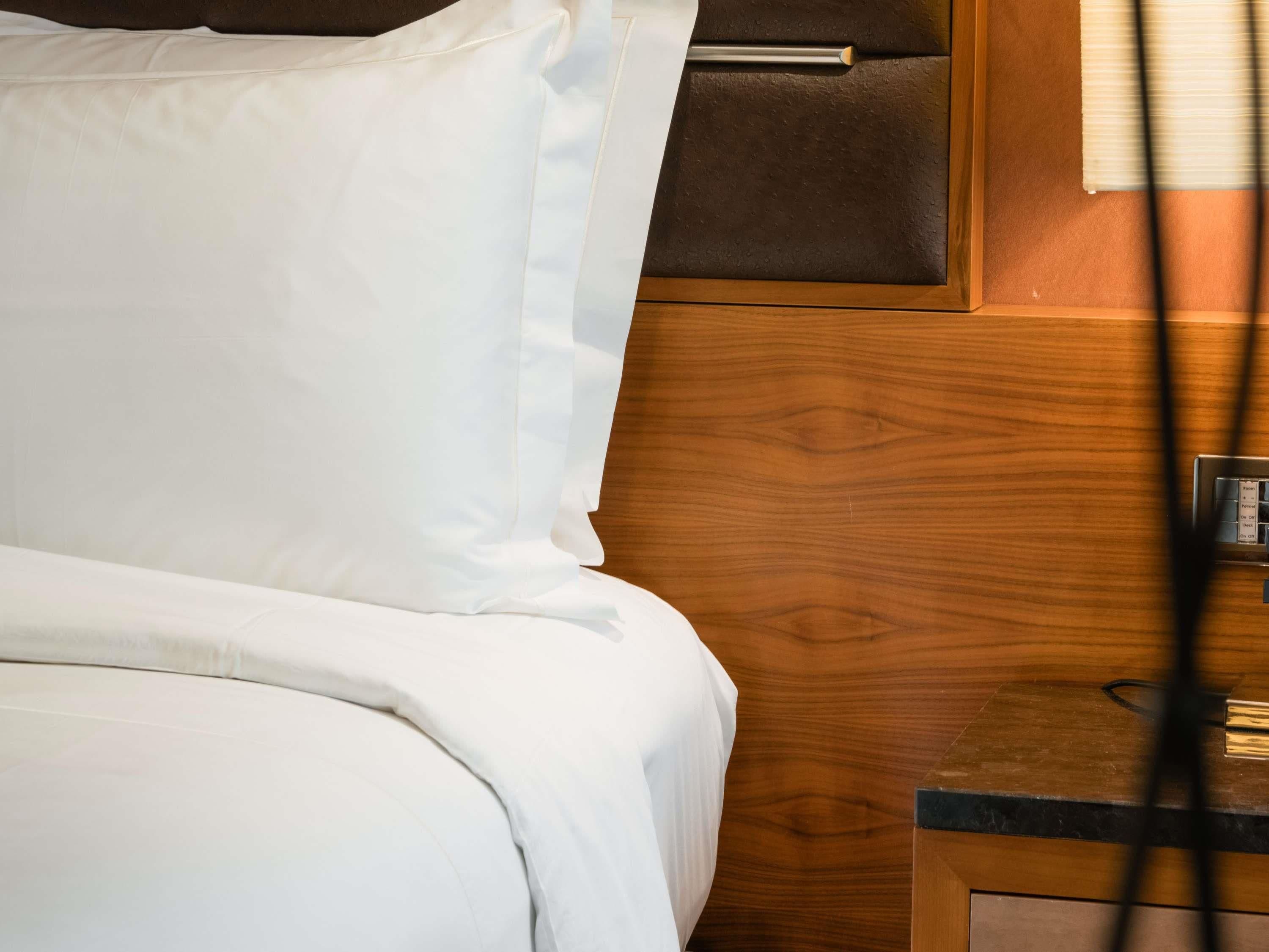 Una cama con ropa de cama blanca, cabecera de madera, mesita de noche con lámpara y pared de paneles de madera.