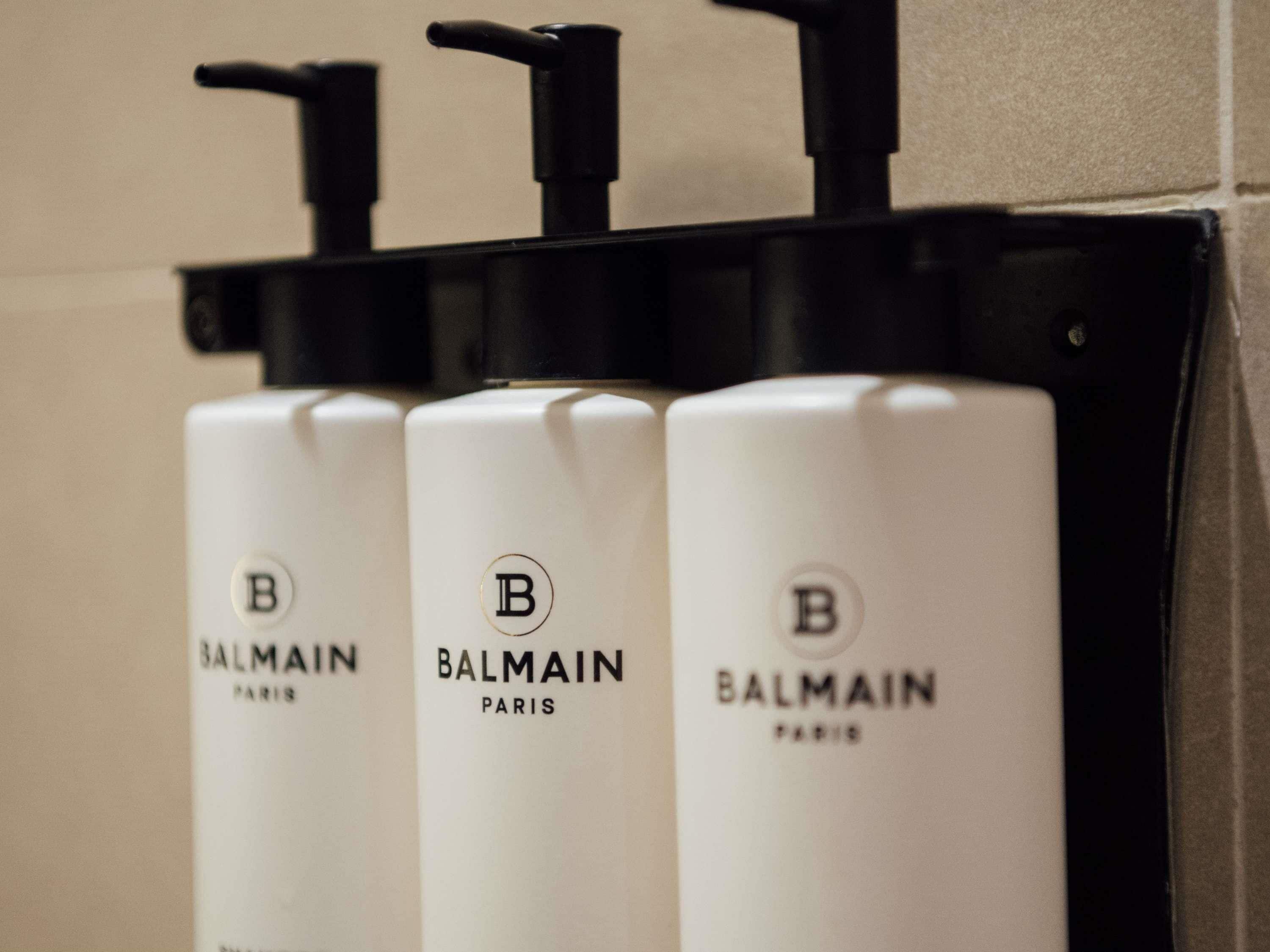 Trois flacons de parfum Balmain Paris, de couleur blanche, sont fixés sur un distributeur mural noir.