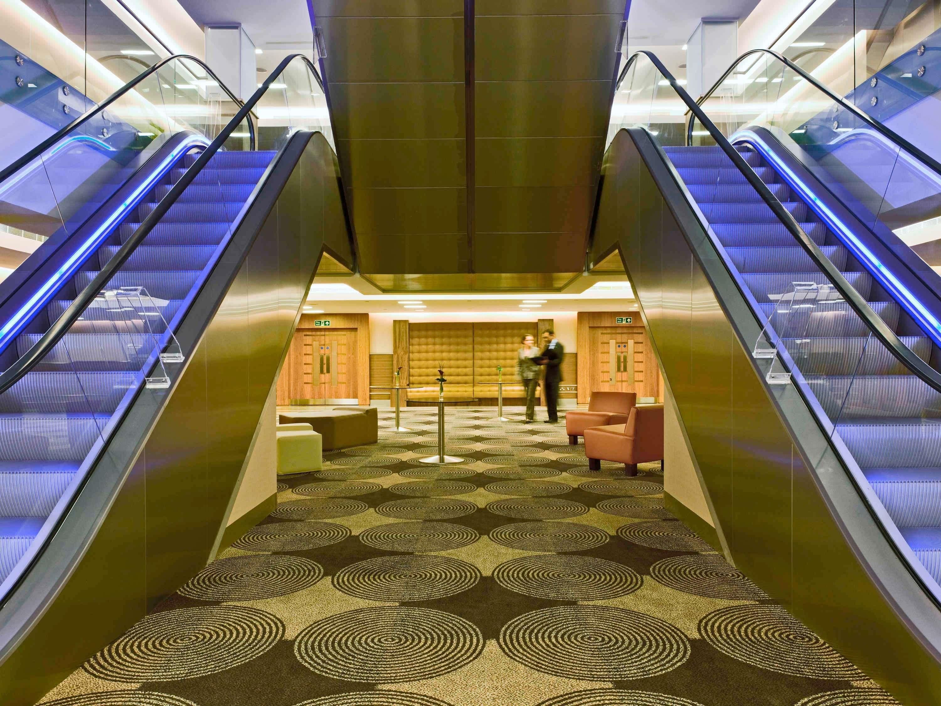 An interior view of a hotel lobby with two escalators, a patterned carpet, seating areas, and a reception desk.