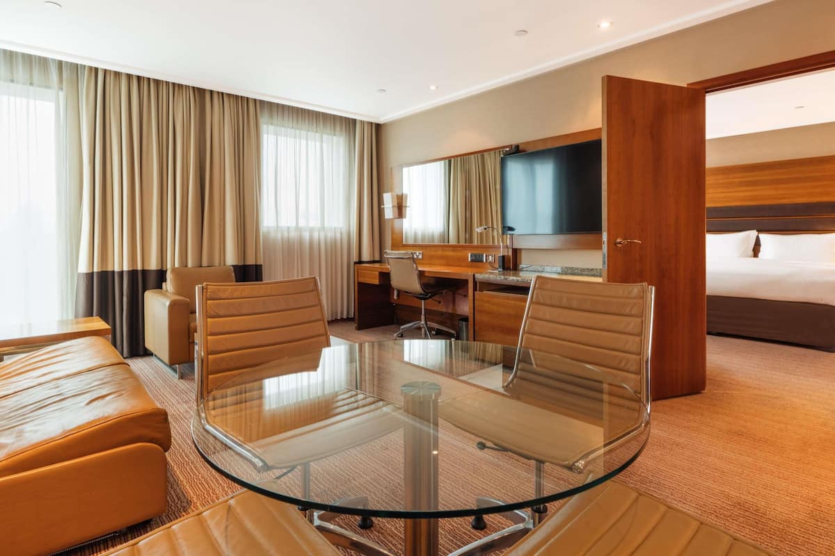 A hotel room with a glass table, leather chairs, a bed, a desk, and a TV.