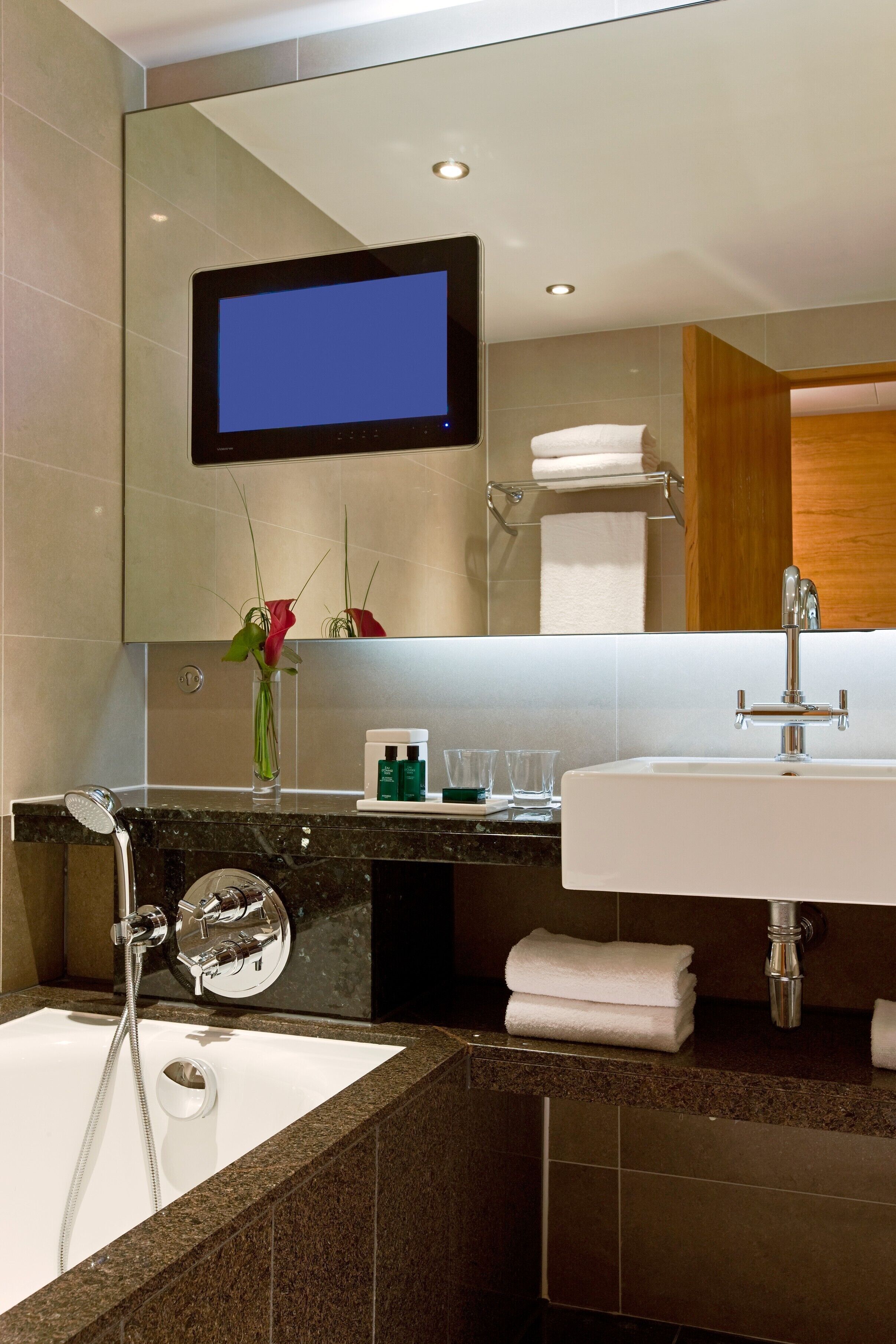 A modern bathroom with a large mirror, a flat-screen TV, a bathtub, and a sink.