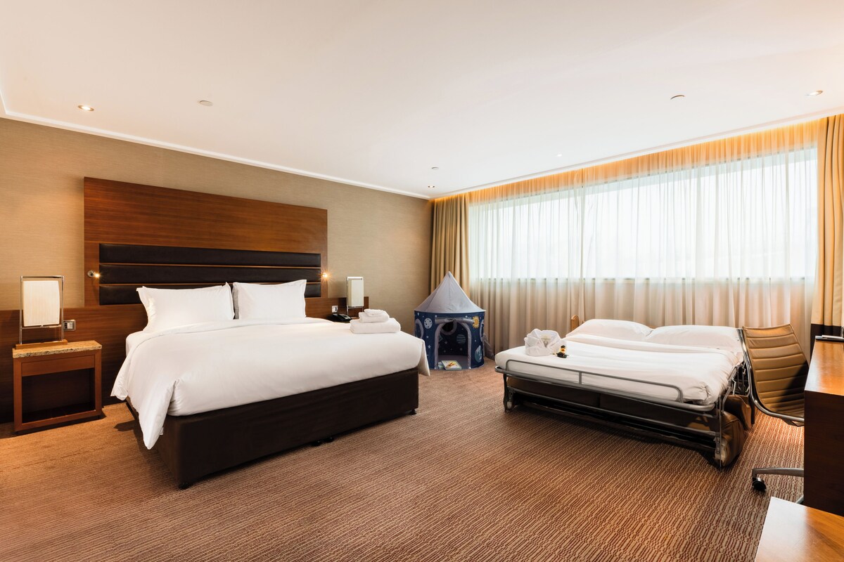A hotel room with a large bed, a desk, a chair, a small play tent, and a large window with curtains.