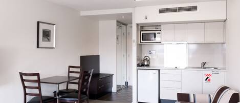 A compact kitchen with white cabinets, a microwave, and a small refrigerator. There is a dining area with a table and chairs, and a kitchenette with a stove and sink.
