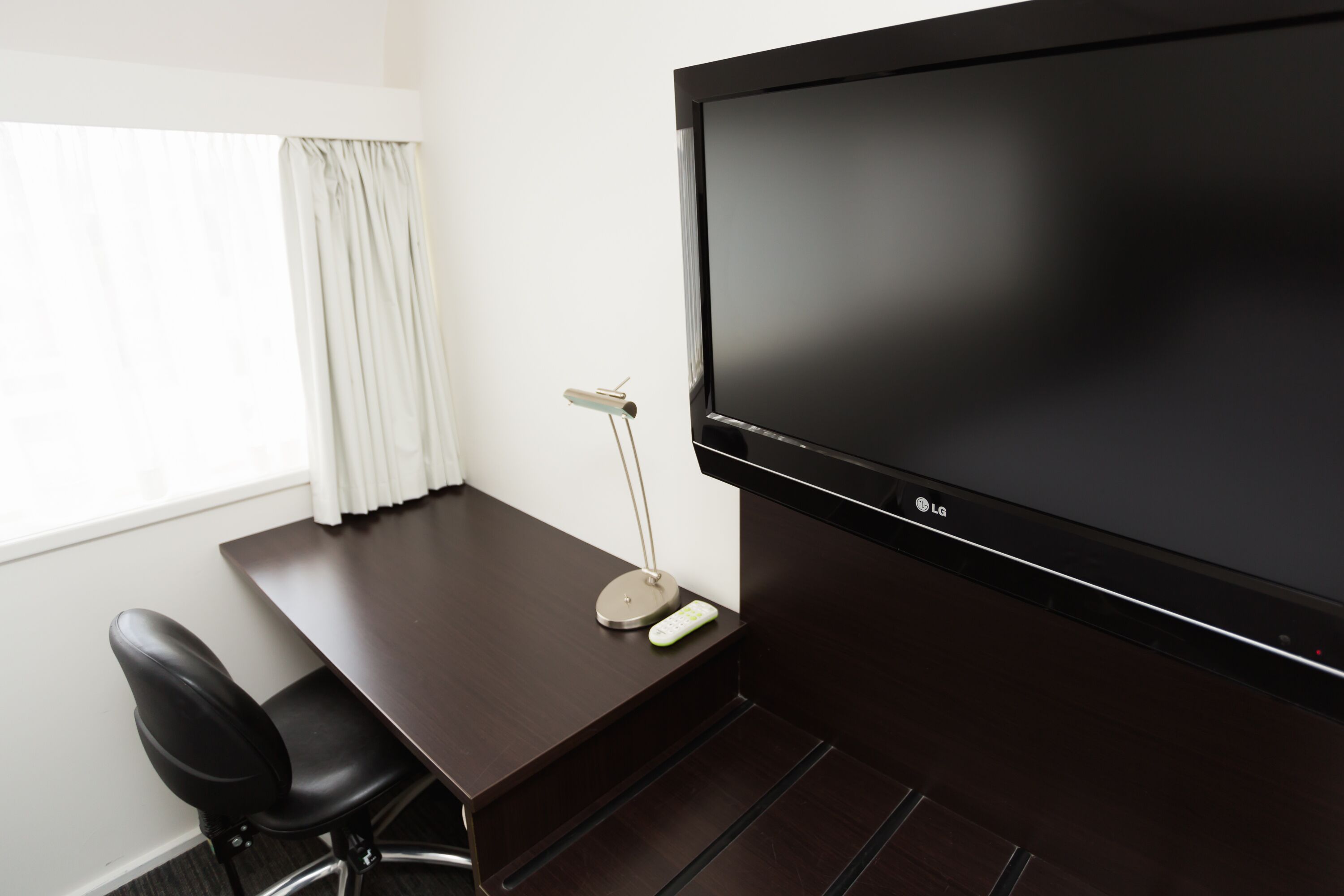 A dark wooden desk with a remote control, a black office chair, and a large flat-screen TV mounted on the wall.