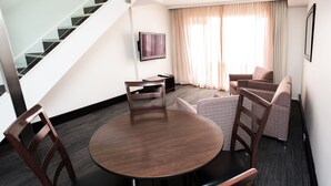 Habitación de hotel con una mesa redonda de madera, cuatro sillas de madera, un sofá, un televisor montado en la pared, una escalera y un gran ventanal con cortinas.