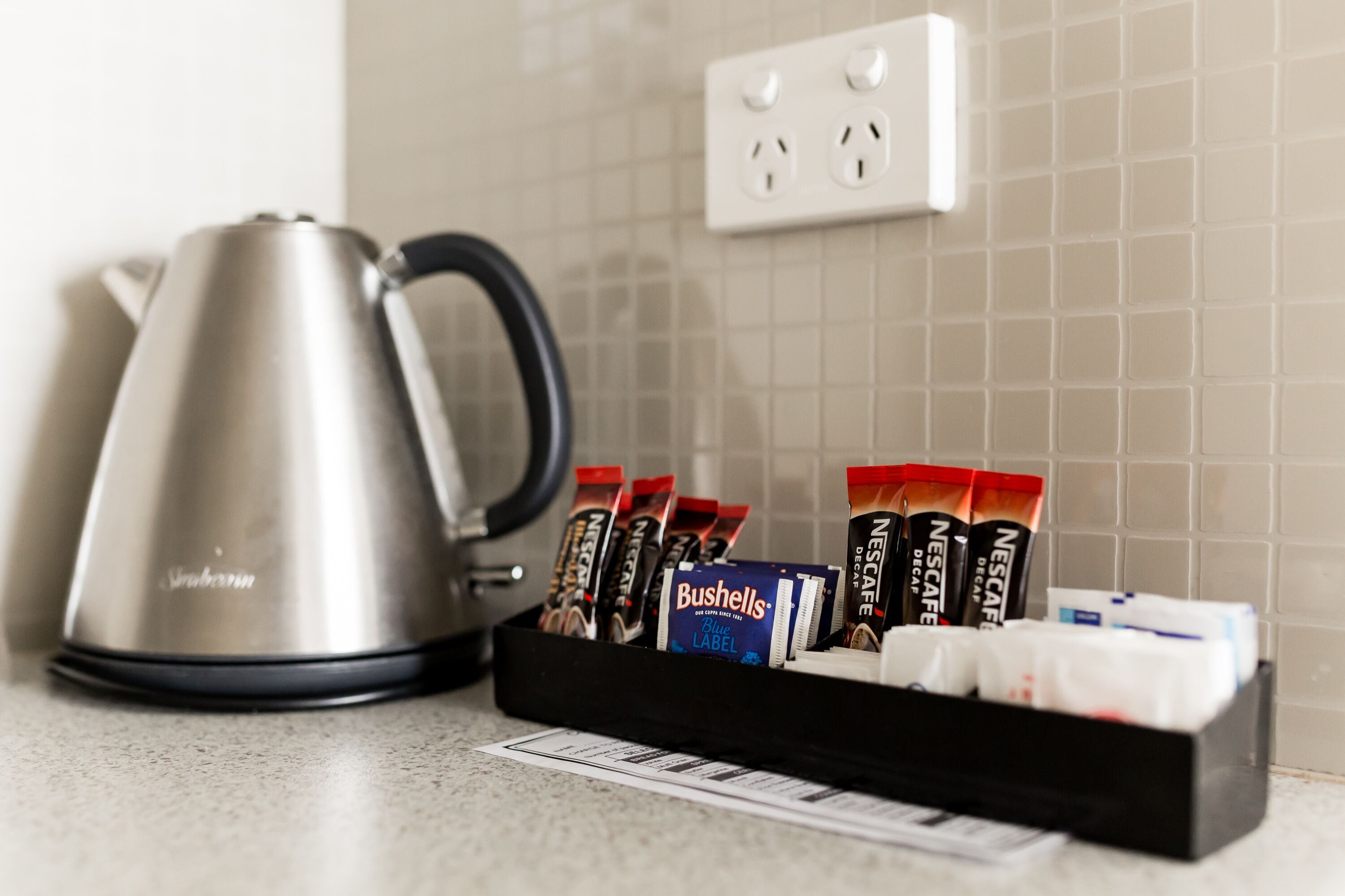 A kitchen counter with a stainless steel kettle, a black tray holding coffee pods, and a white electrical outlet.