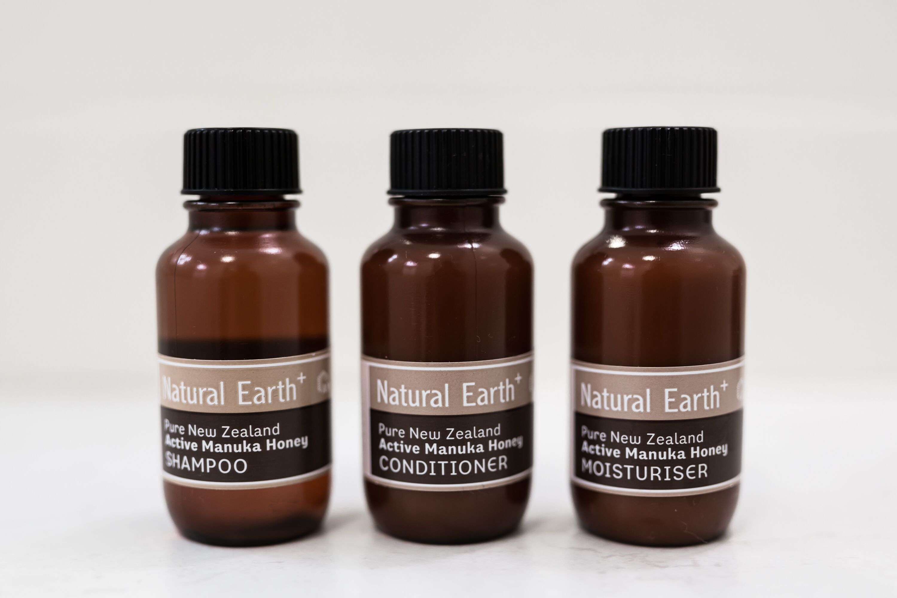 Three brown bottles labeled 'Natural Earth+ Pure New Zealand Active Manuka Honey' with different product names: SHAMPOO, CONDITIONER, and MOISTURISER.