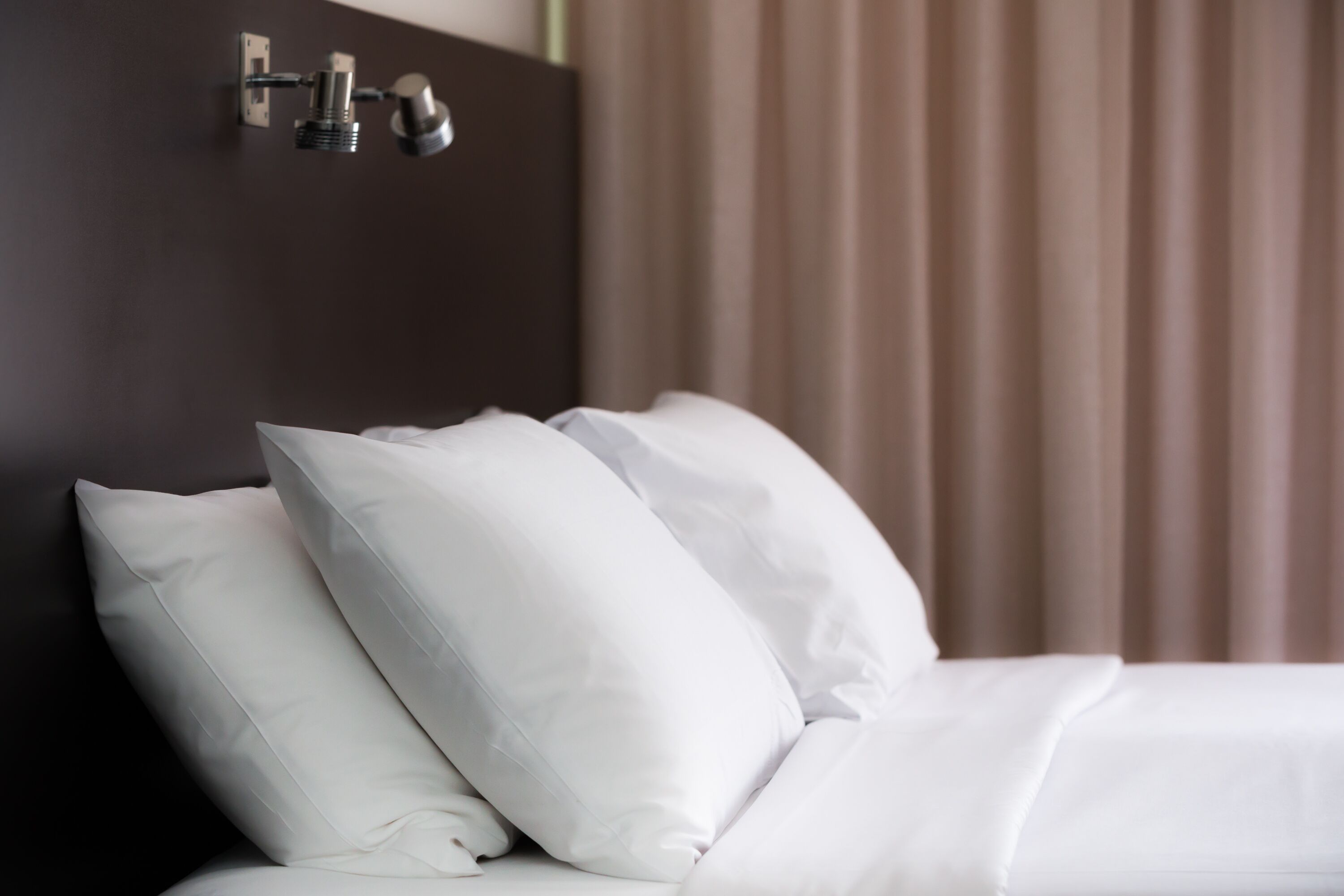 A neatly made bed with white bedding and pillows, a dark headboard, and a wall-mounted light fixture.