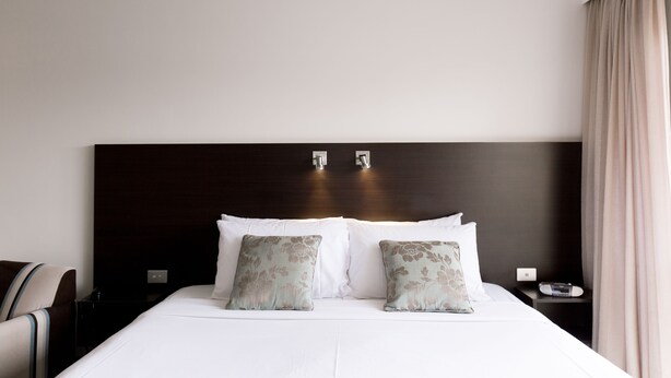 Habitación de hotel con una cama grande, dos almohadas, una mesita de noche, una silla y un ventanal con cortinas.
