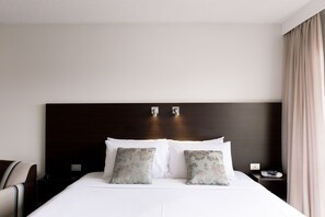 Ein Hotelzimmer mit einem großen Bett, zwei Kissen, einem Nachttisch, einem Sessel und einem Fenster mit Vorhängen.