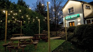Una mesa de picnic de madera y bancos en un jardín con luces colgantes. Se ve un edificio en el fondo con un letrero que dice "WEE WAIF".