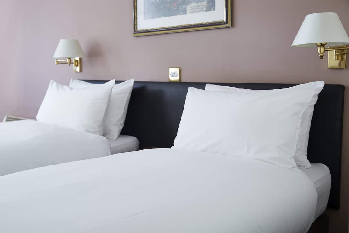 A hotel room with two beds, white bedding, a dark headboard, and wall-mounted lamps.