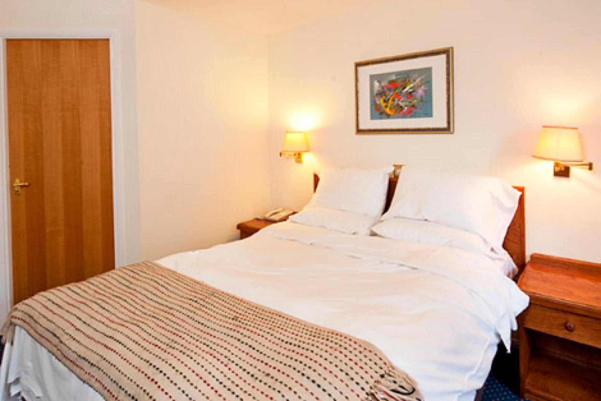 Ein Hotelzimmer mit Bett, Nachttisch, Lampe und einem Gemälde an der Wand.