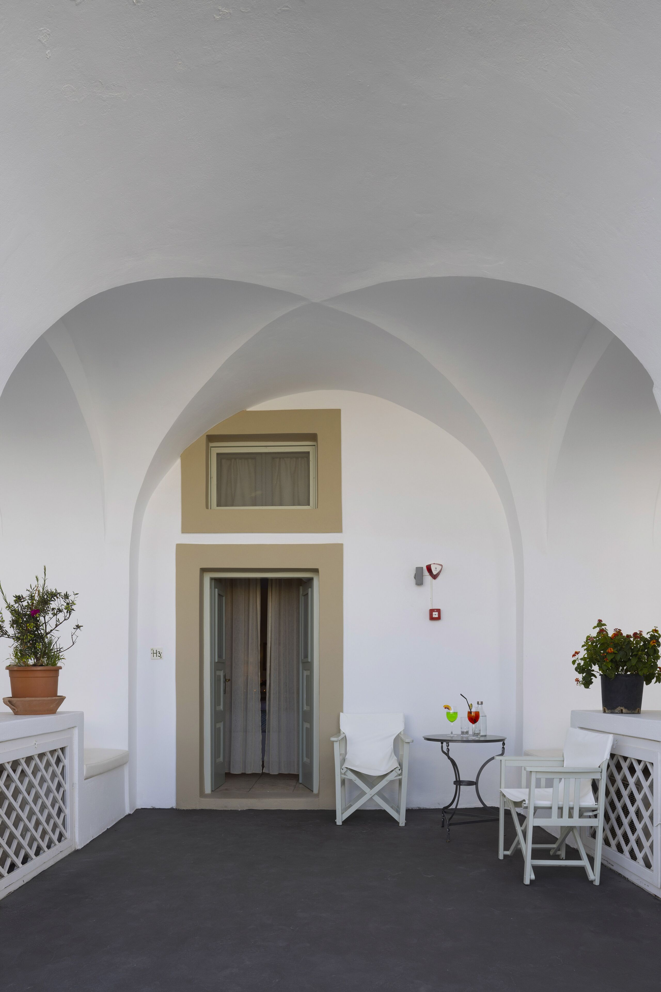 Une pièce aux murs blancs, dotée d’un haut plafond en arc de cercle, d’une porte, et de deux chaises blanches, ainsi que d’une petite table et de plantes en pot.