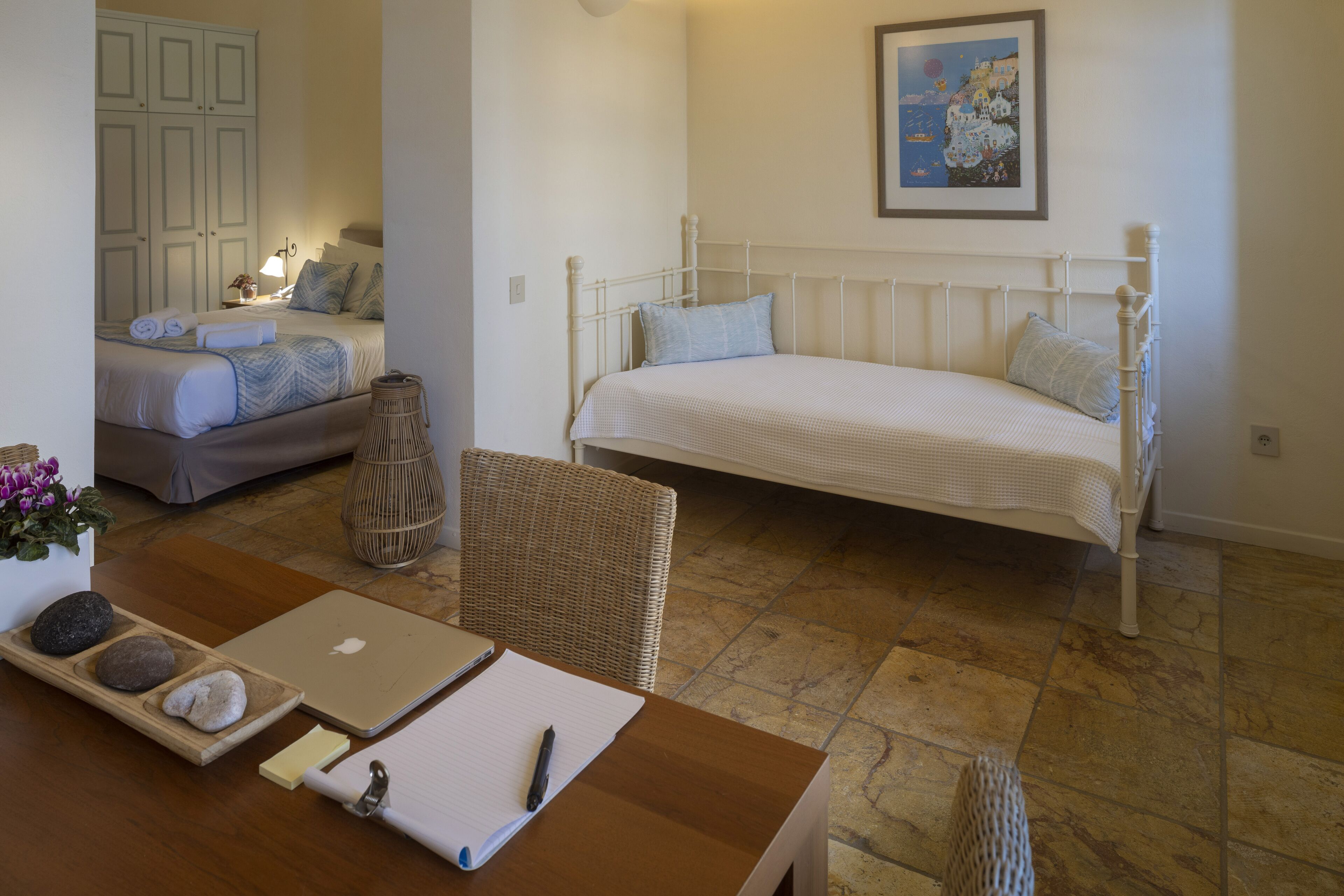 Un dormitorio con cama, un escritorio con computadora portátil, una silla de mimbre y una mesita de noche con lámpara.