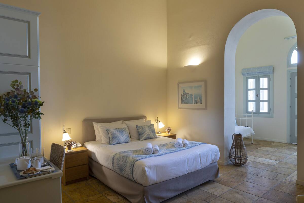 Quarto de hotel com uma cama grande, mesa de cabeceira com abajur, vaso com flores e um quadro na parede.