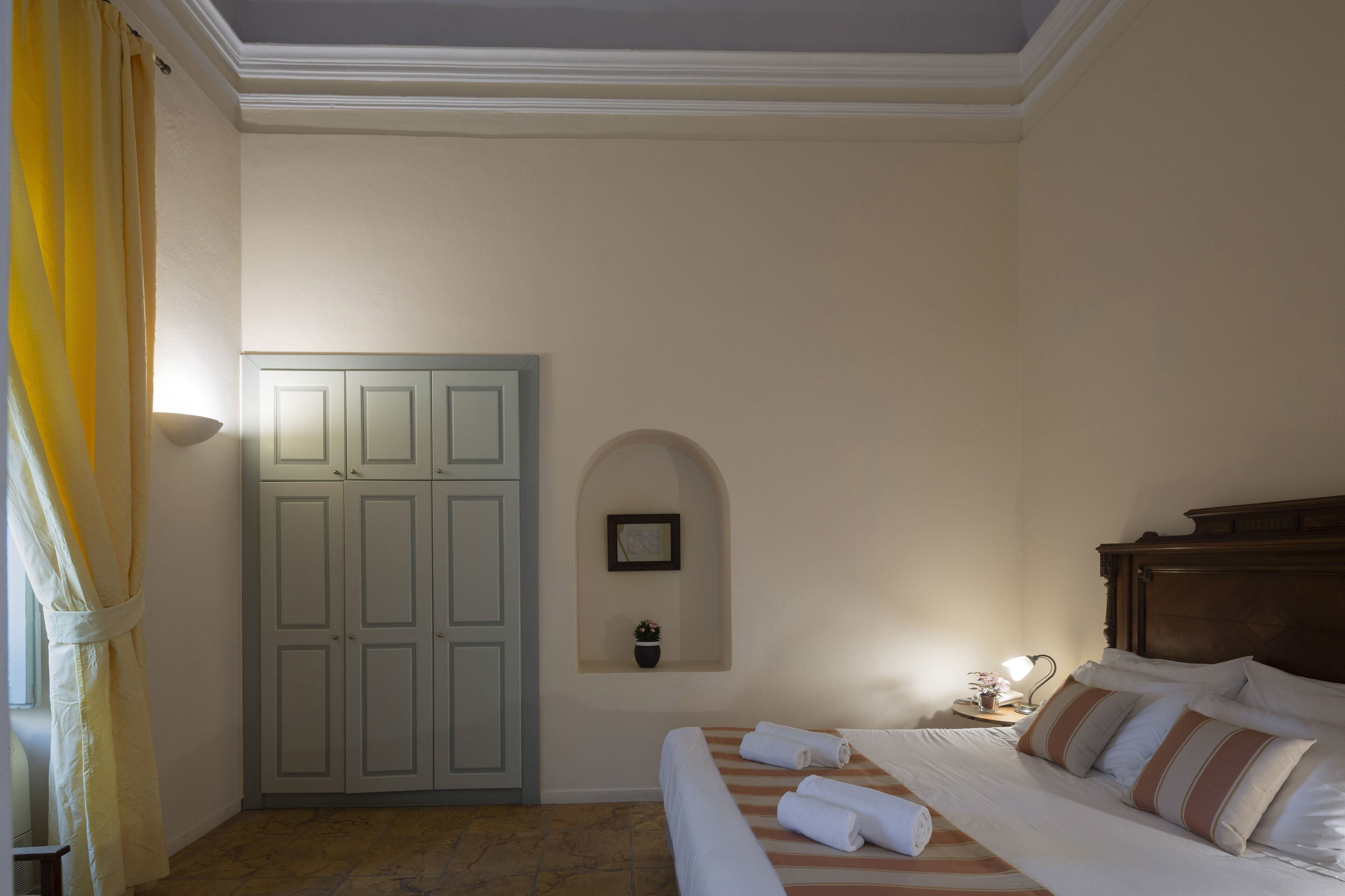 Une chambre à coucher comprenant un lit, une armoire, une petite étagère fixée au mur avec une plante, et une fenêtre avec des rideaux.