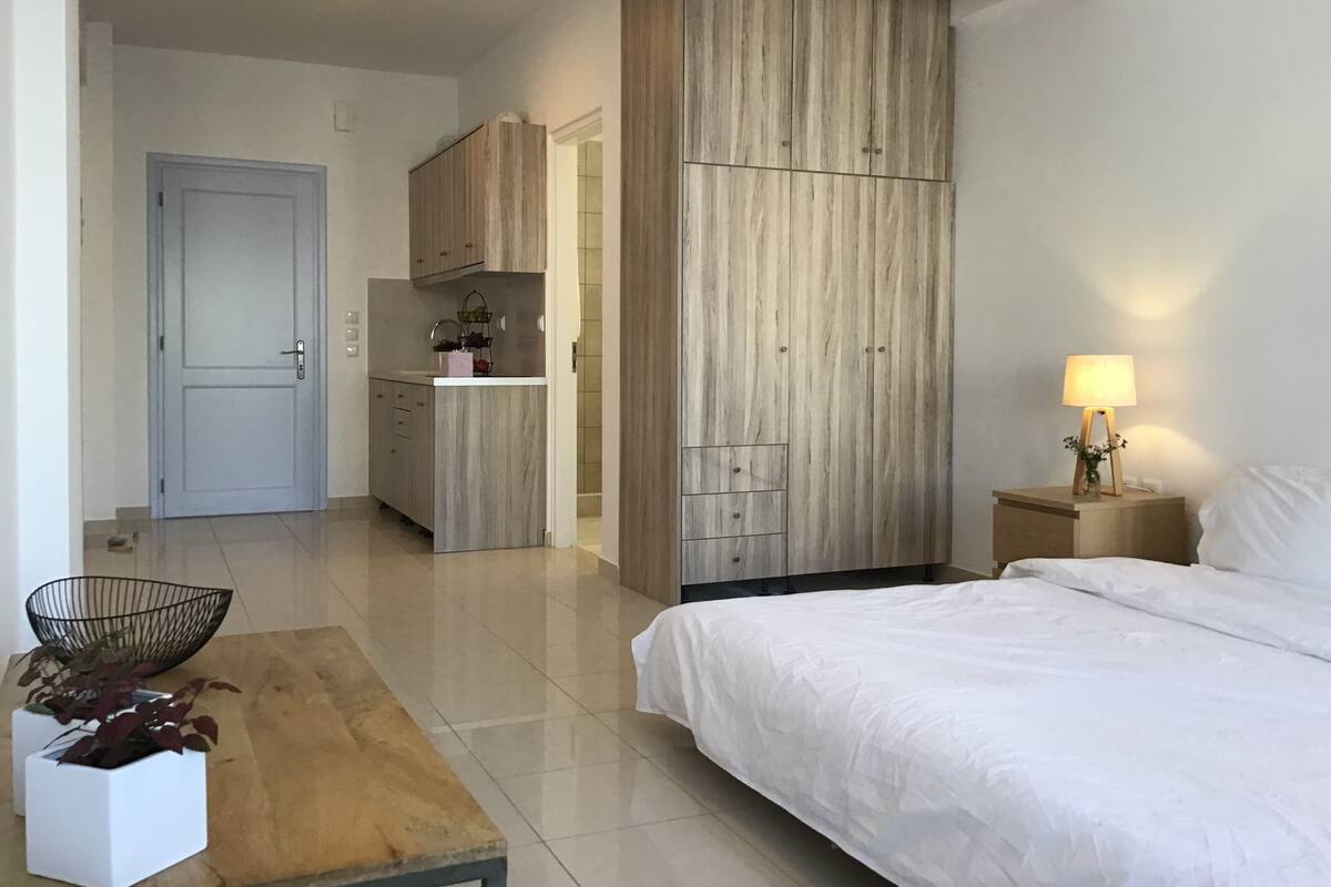 Quarto com cama, mesa de cabeceira com abajur, guarda-roupa de madeira e pequena cozinha com armários e pia.