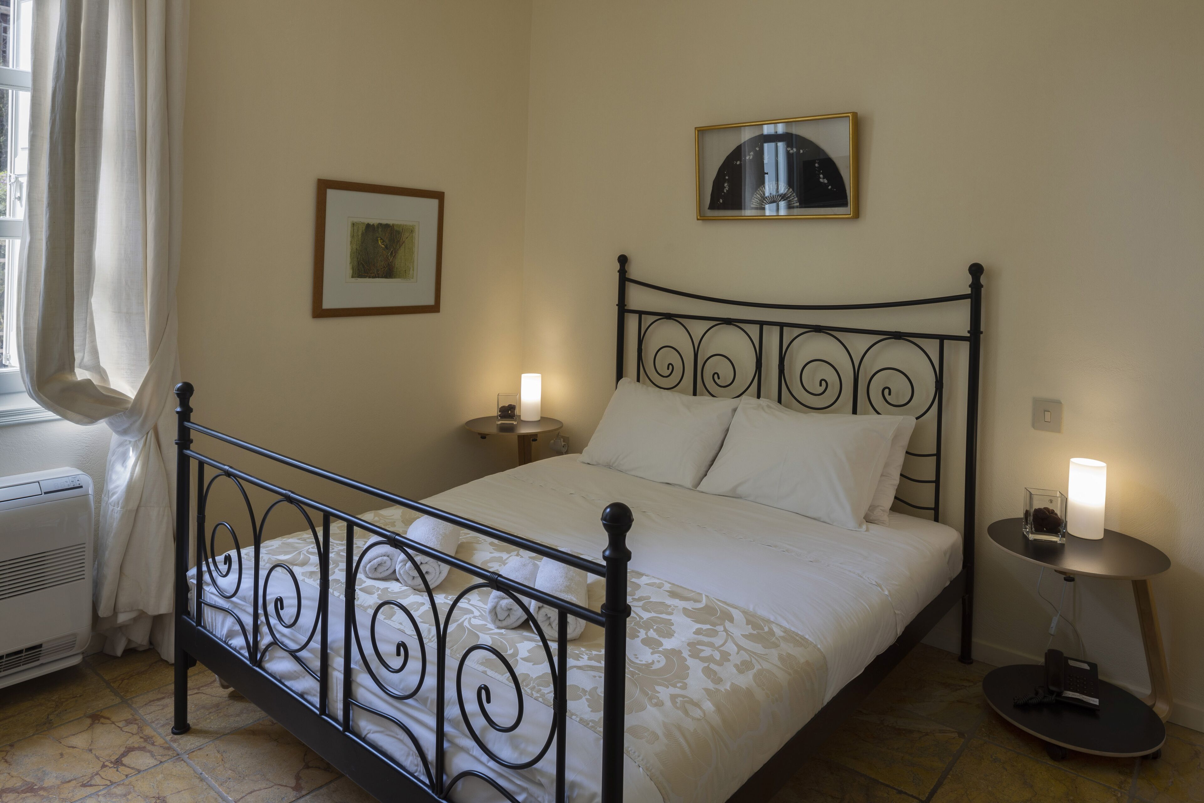 A neatly made bed with a metal frame, two bedside tables with lamps, a framed picture on the wall, and a window with curtains.
