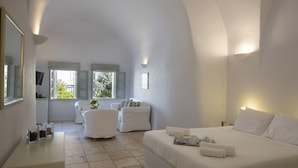 Una habitación de hotel moderna con una cama grande, una zona de estar con sofá y sillones, una mesita con un jarrón de flores y un televisor sobre un mueble.