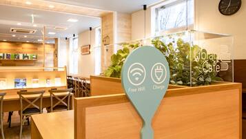 Un café moderno en una biblioteca con mobiliario de madera, una planta verde y un cartel que indica wifi gratuito y puntos de carga.