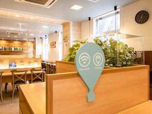 A modern library cafe with wooden furniture, a green plant, and a sign indicating free wifi and charging. - Comfort Hotel Saga (Saga)
