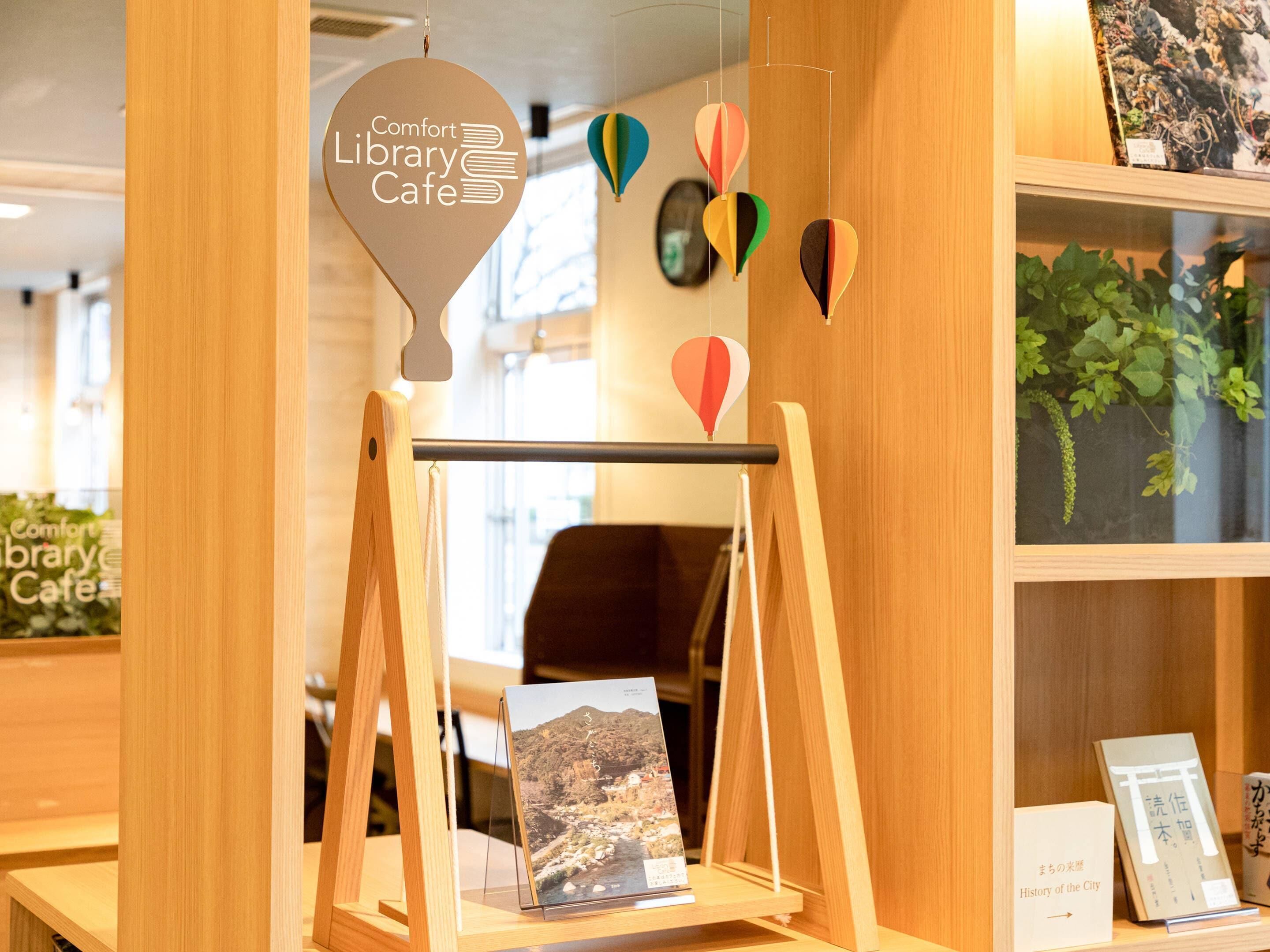 Un café biblioteca con un letrero que dice "Comfort Library Cafe", una exhibición de libros y vistas de un paisaje montañoso a través de una ventana.