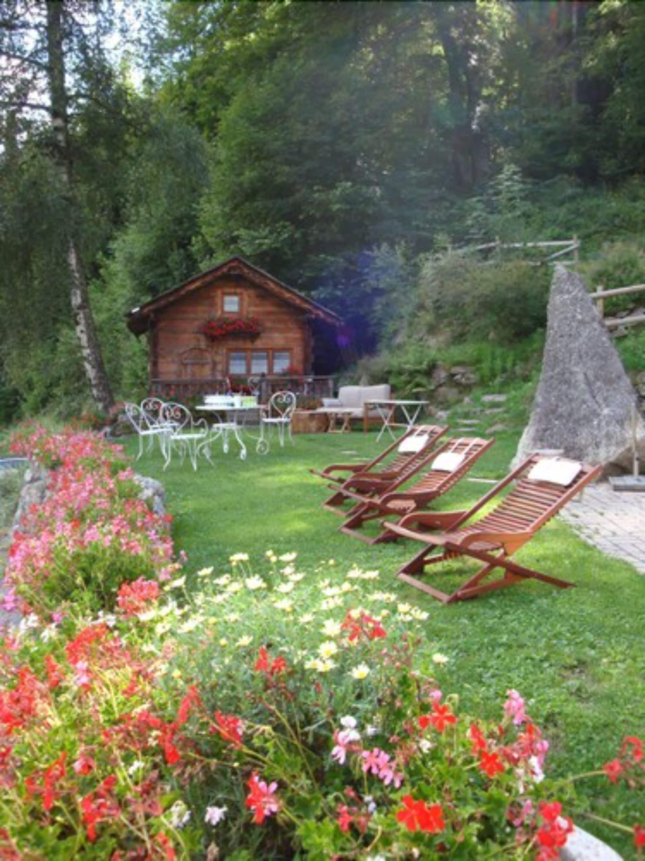 Une cabane en bois dotée d’un balcon, entourée d’un jardin luxuriant agrémenté de fleurs rouges et d’une structure en pierre.