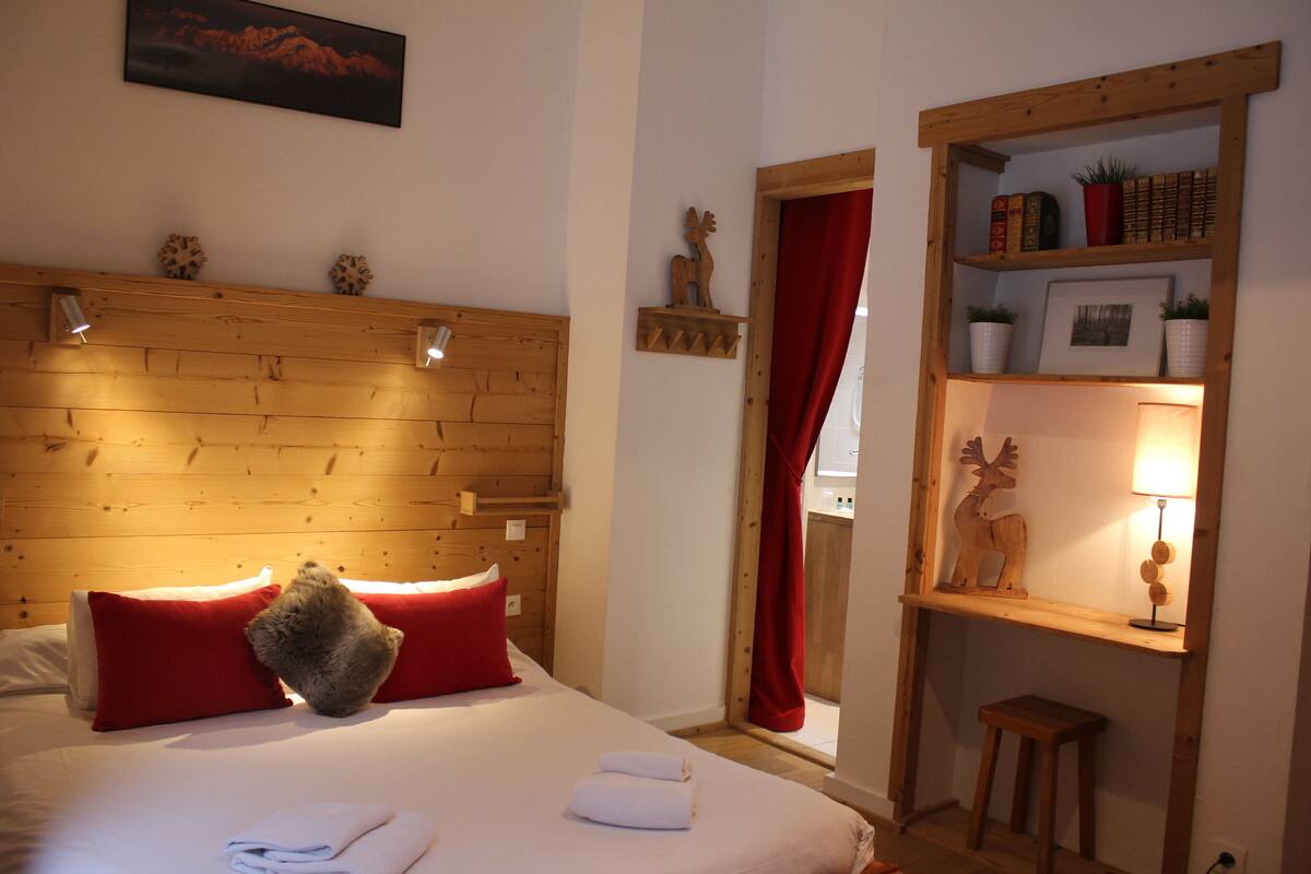 A room with a wooden headboard, red pillows, a bed with white linens, a small wooden stool, and a shelf with books and decorative items.