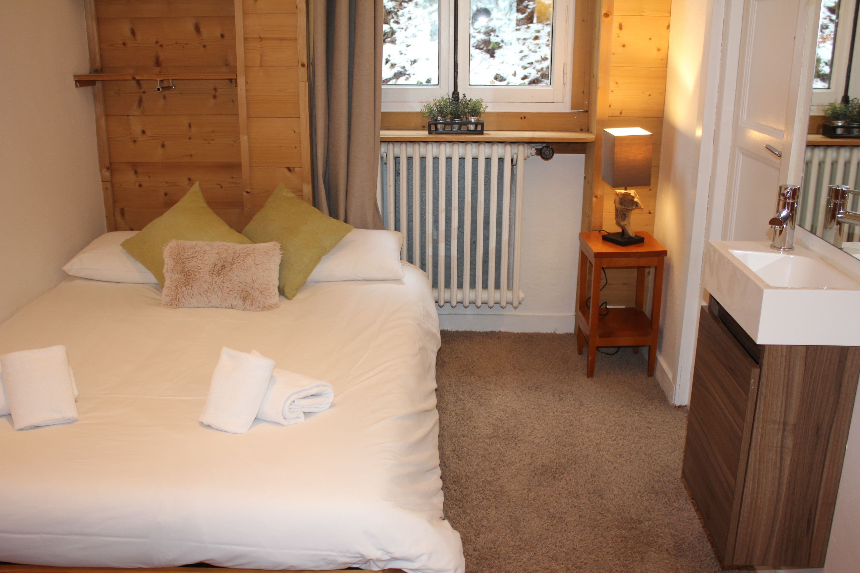 Une chambre à coucher avec un lit, des oreillers, une table de chevet avec une lampe, un radiateur et une fenêtre donnant sur un paysage enneigé.