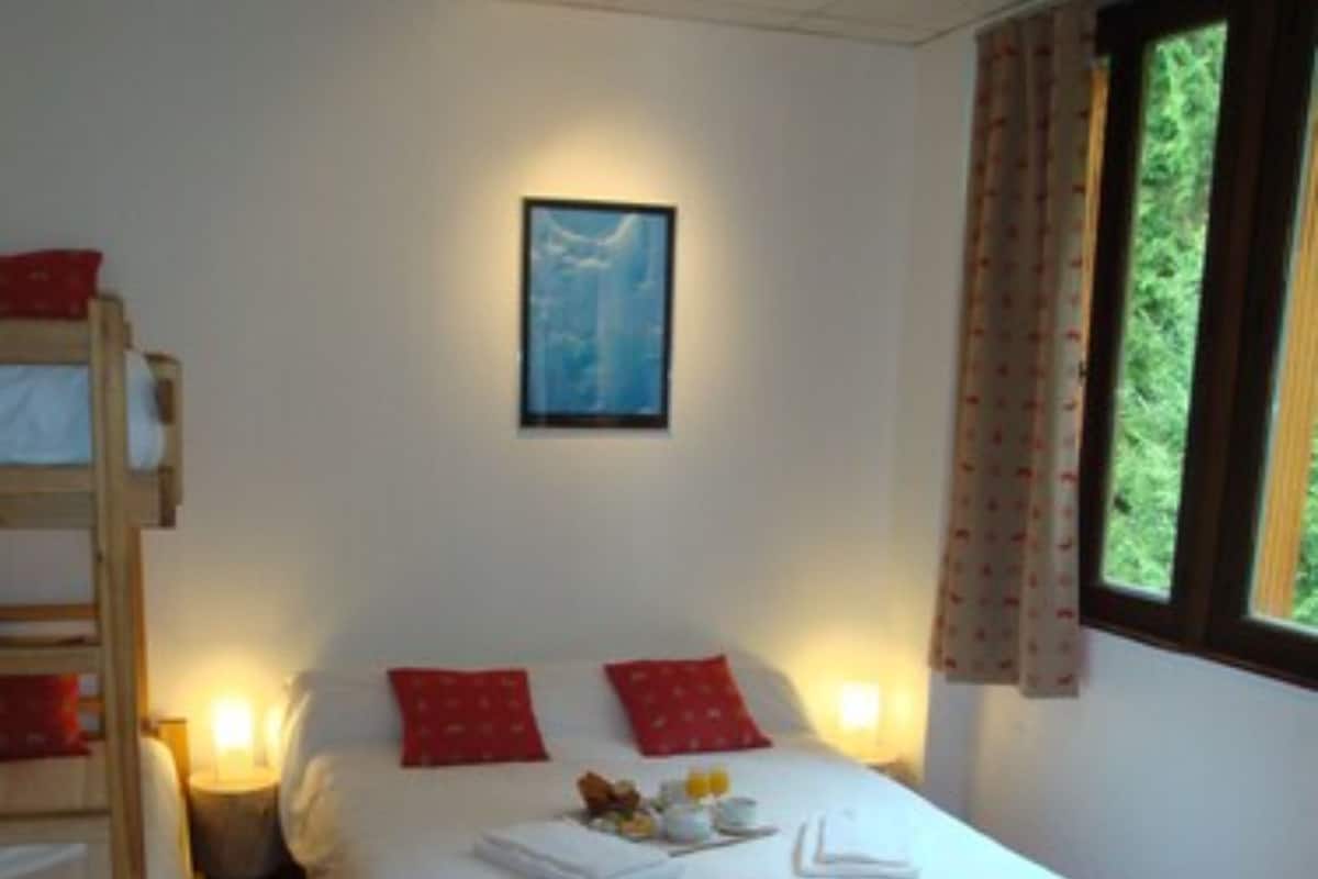 A hotel room with a single bed, red pillows, a wooden bunk bed, a framed picture on the wall, and a window with curtains.