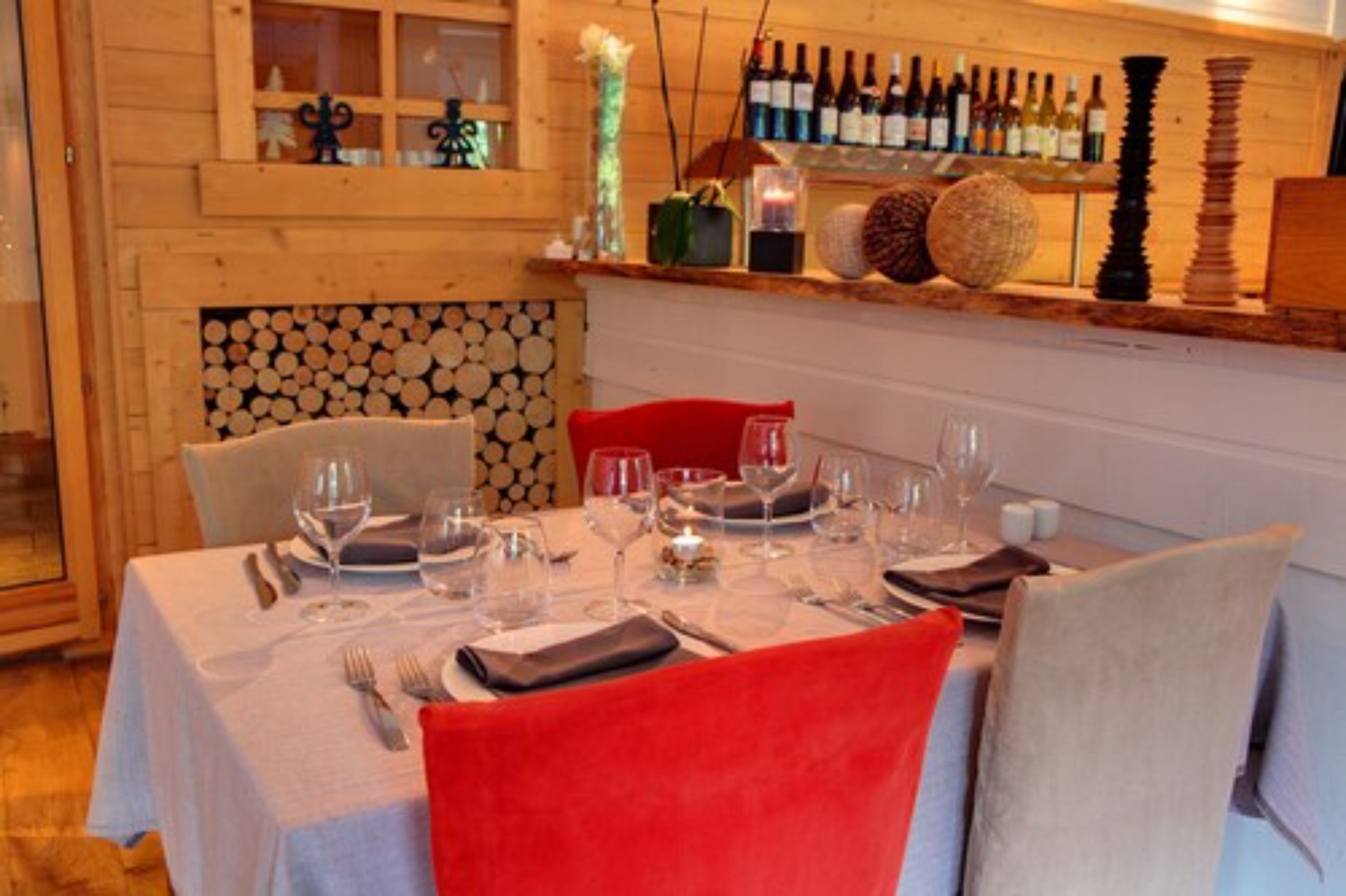 A dining area with a table set for four, a wooden wall with a log display, and a shelf with wine bottles.