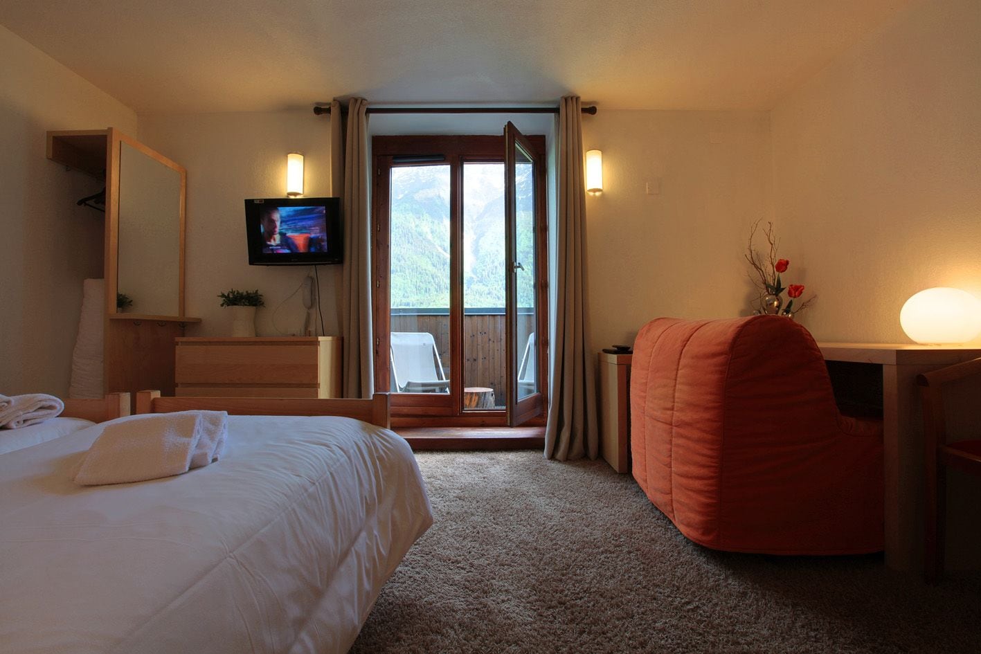 A hotel room with a bed, a desk, a TV, and a balcony with a view of the outdoors.