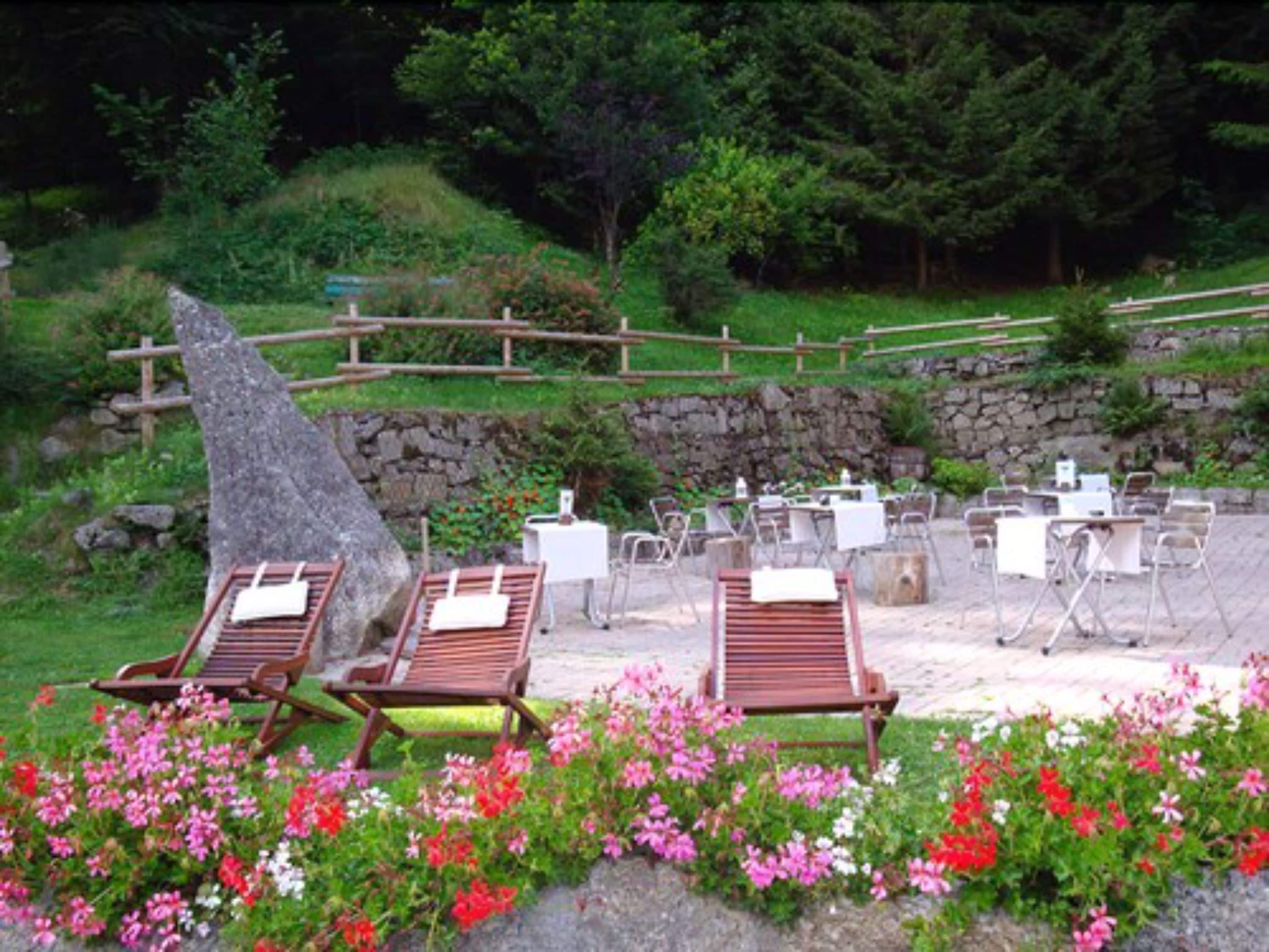 An outdoor seating area with wooden lounge chairs and tables, surrounded by colorful flowers and a stone wall.