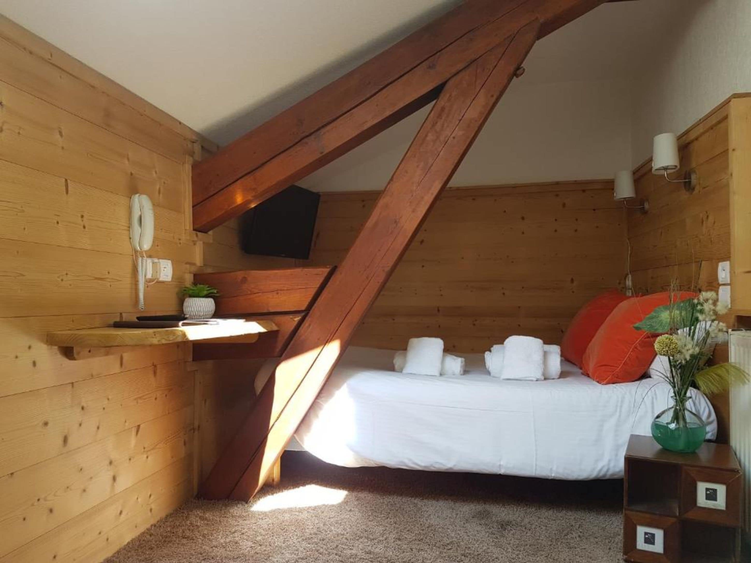 Una camera da letto accogliente in legno con soffitto spiovente, un letto con lenzuola bianche e un cuscino rosso, un comodino con una lampada e un televisore montato a parete.