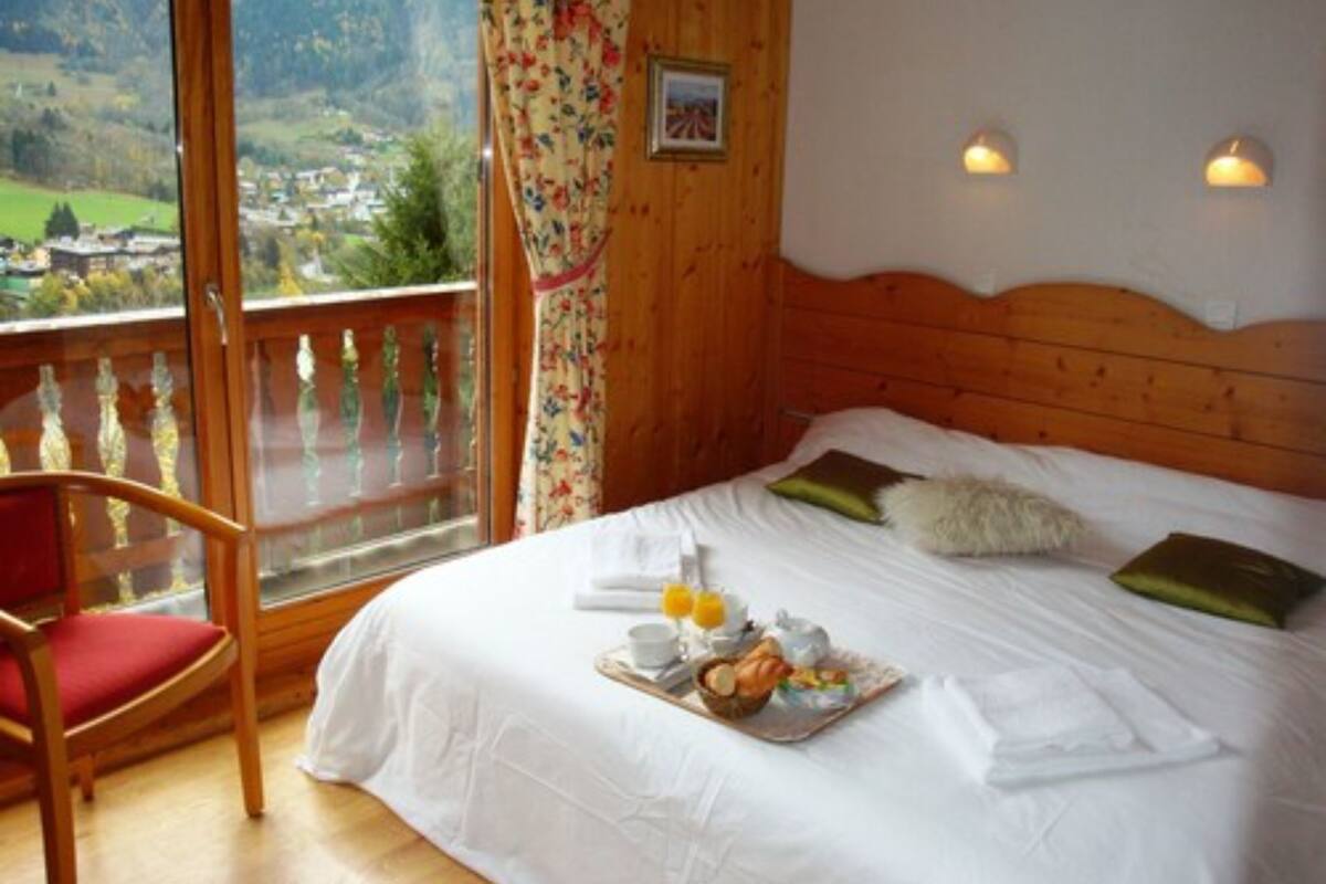 A bedroom with a wooden headboard, a bed with white linens, a breakfast tray, a chair, and a balcony with a view of a village.