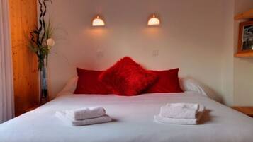 Una camera d'albergo con un letto, cuscini rossi, due luci a parete e un vaso con una pianta.