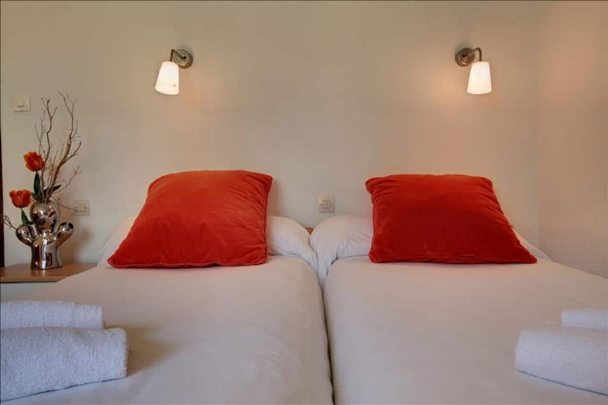 A hotel room with two beds, each with a red pillow and white bedding. There are wall-mounted light fixtures above the beds.