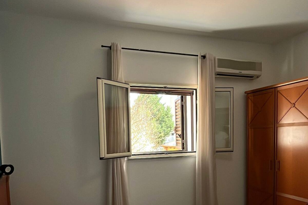 A bedroom with a bed, a window with curtains, a wardrobe, and an air conditioning unit.