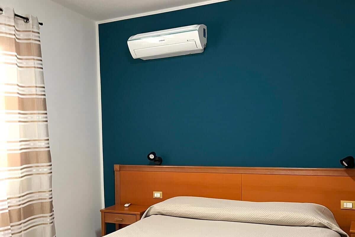 A hotel room with a bed, a bench, a nightstand, and an air conditioning unit on the wall.