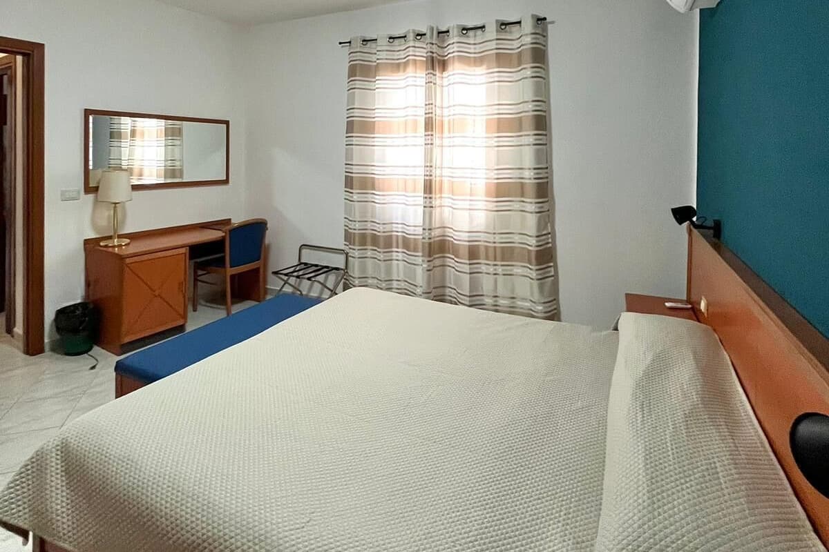 A hotel room with a bed, a desk, a chair, a mirror, and a window with curtains.