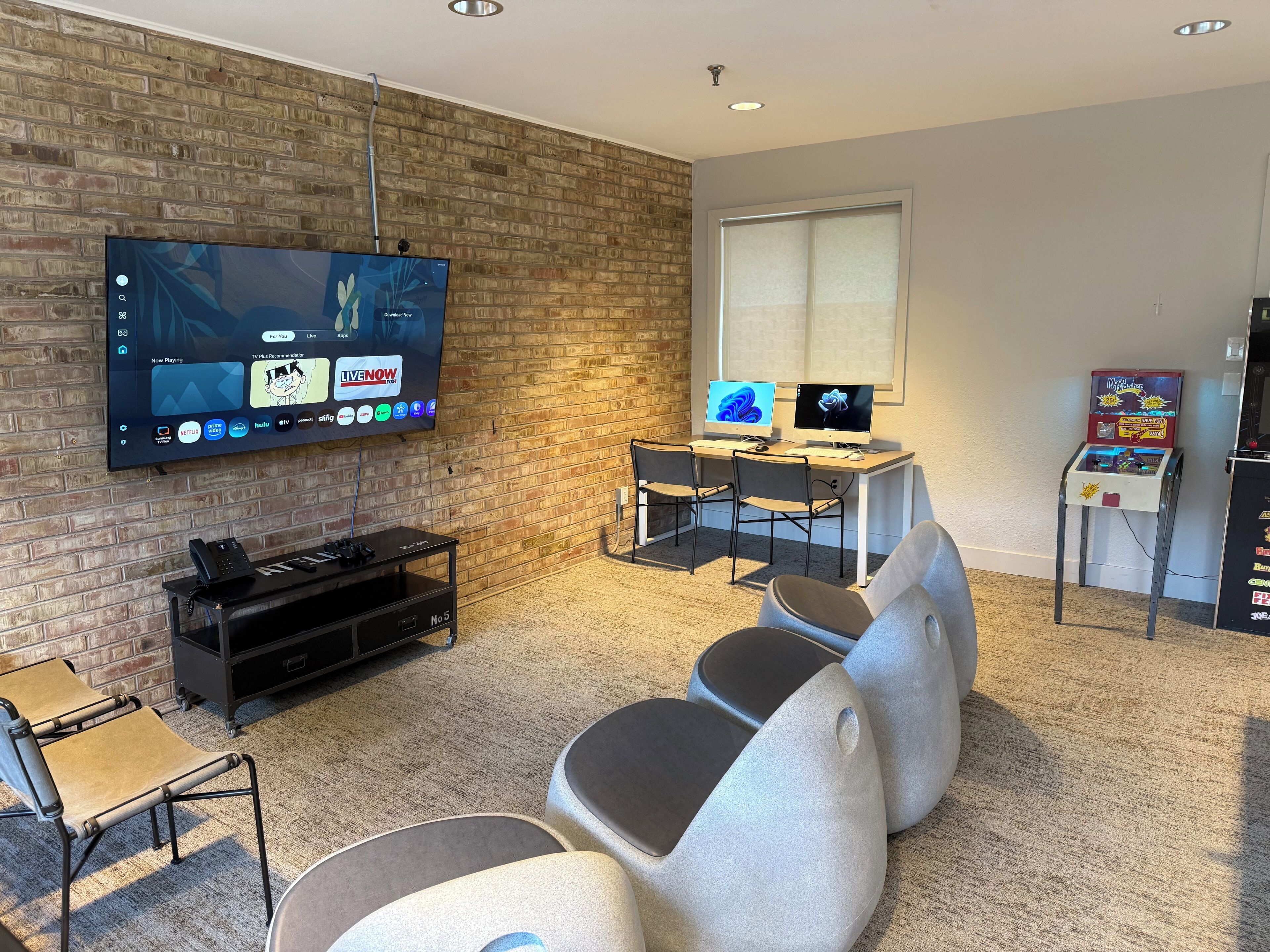 Ein Wohnzimmer mit Fernseher, zwei Monitoren, einer Spielkonsole und einem kleinen Tisch mit einem Spiel.