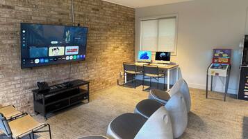 Ein Wohnzimmer mit Fernseher, zwei Monitoren, einer Spielkonsole und einem kleinen Tisch mit einem Spiel.