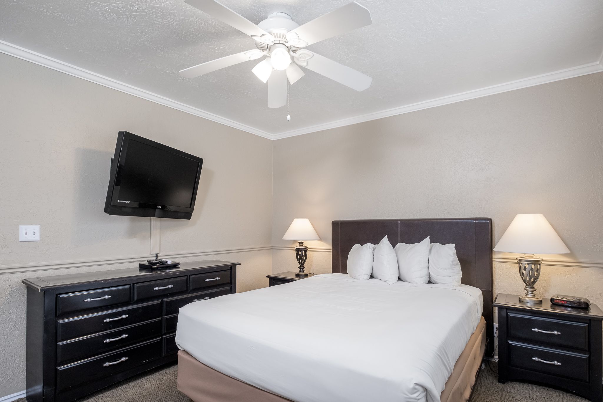 A hotel room with a bed, two bedside tables with lamps, a flat-screen TV mounted on the wall, and a ceiling fan.